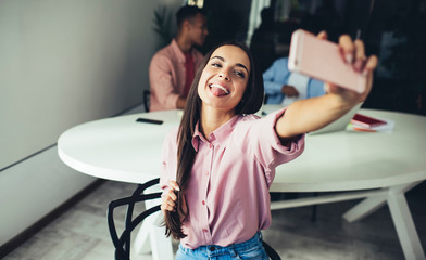 Positive blogger posing for cellular camera while making selfie and showing tongue, Caucasian female fooling during taking photo on application on modern smartphone gadget connected to 4g wireless