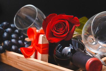 Obraz na płótnie Canvas red rose, bottle of wine, 2 glasses, dark grapes, corkscrew, gift box with ribbon on black background
