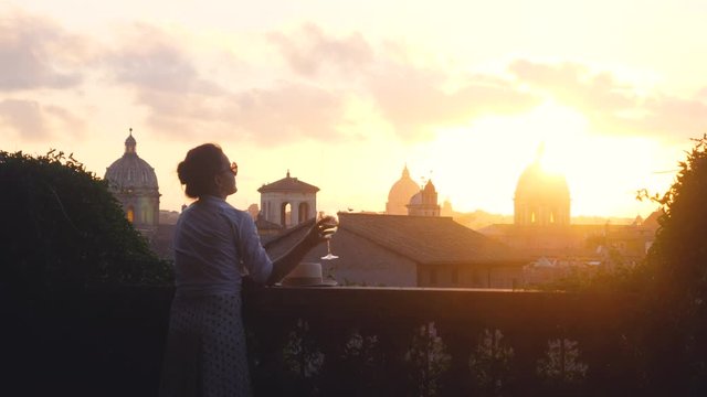 Young woman tourist fashion white dress with glass of white wine in front of panoramic view of Rome cityscape from campidoglio terrace at sunset. Landmarks, domes of Rome, Italy.