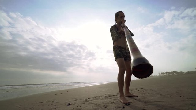 Young musician man playing on didgeridoo on beach at sunset.
