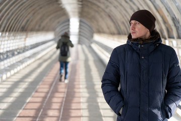 A young man in a winter jacket stands in a light tunnel and looks away. Profile view of handsome young man outdoor, looking away thinking