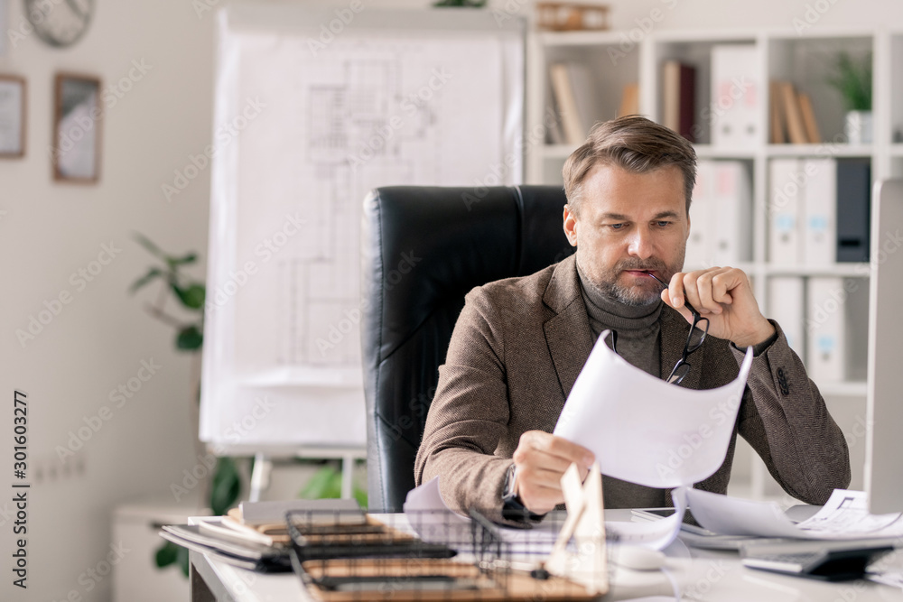 Poster Pensive mature architect looking at sketch on paper while sitting in armchair