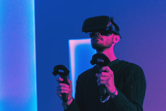 An adult guy in VR glasses with a gamepad in his hands plays a game in a room lit by neon light
