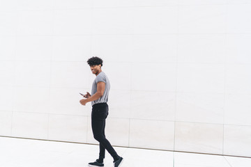 Cheerful handsome young curly man in casual outfit surfing mobile while walking on marble passage