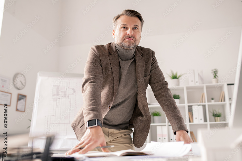 Poster Pensive mature architect leaning against desk with papers while planning work