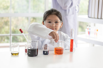 A 6 year old Asian girl wearing a white scientist uniform Learning And conducted a scientific experiment on a white table with a measuring cup and test tube placed, to education concept.