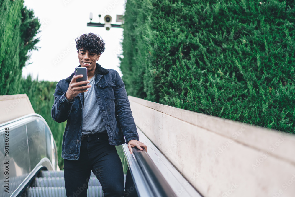 Poster happy hipster guy going down at city escalator and using modern cellphone gadget for online communic