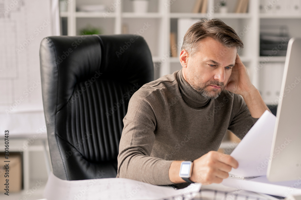 Poster Mature engineer with papers looking at one of sketches while checking them