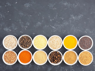 Buckwheat, millet, allspice, fennel, oat, pepper chili, masala, fenugreek, cloves, turmeric, cumin, mustard seeds, green cardamom in white bowls on black concrete background with copy space