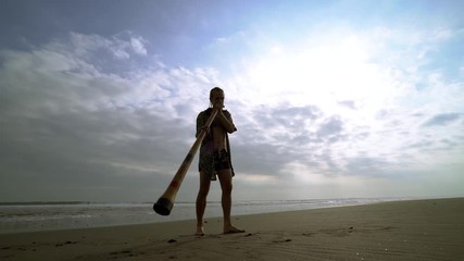Young musician man playing on didgeridoo and girl with guitar on beach at sunset.