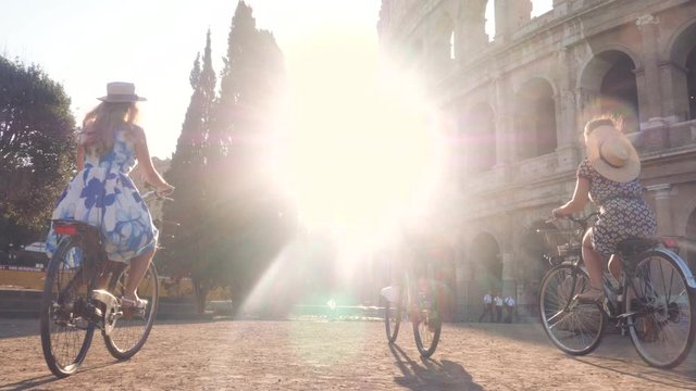 Three happy young women friends tourists riding bikes at Colosseum in Rome, Italy at sunrise.