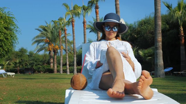 Young woman in hat and sunglasses writes, draws and browsing news feed on a tablet while sitting on a deck chair with coconut water cocktail. Palm trees are at the background.
