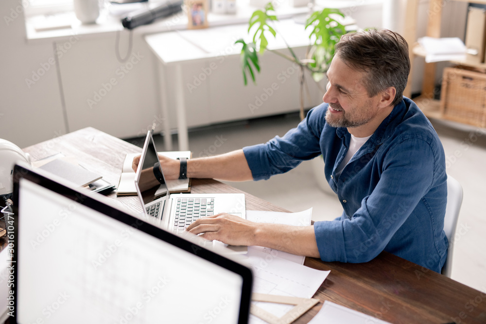 Poster Happy engineer sitting in front of laptop, surfing in the net and making notes