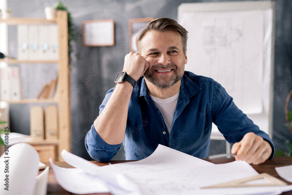 Poster Smiling mature engineer sitting by table while working over new sketches