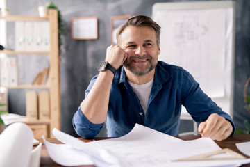 Smiling mature engineer sitting by table while working over new sketches