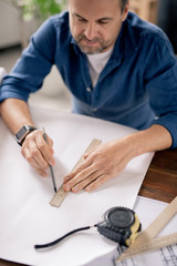 Modern engineer with pencil and ruler drawing line on blueprint by table