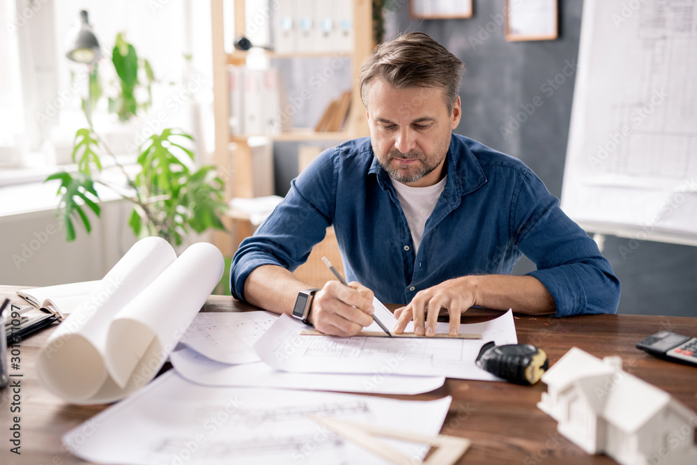 Aufkleber serious architect in casualwear sitting by table while working over new project