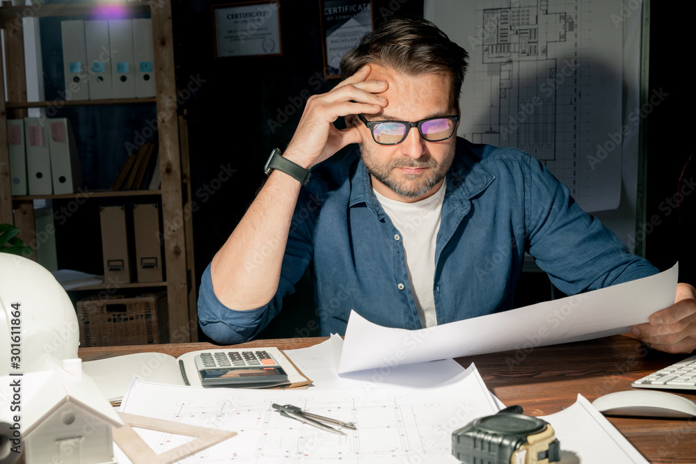 Poster serious mature engineer in eyeglasses checking sketch while working late