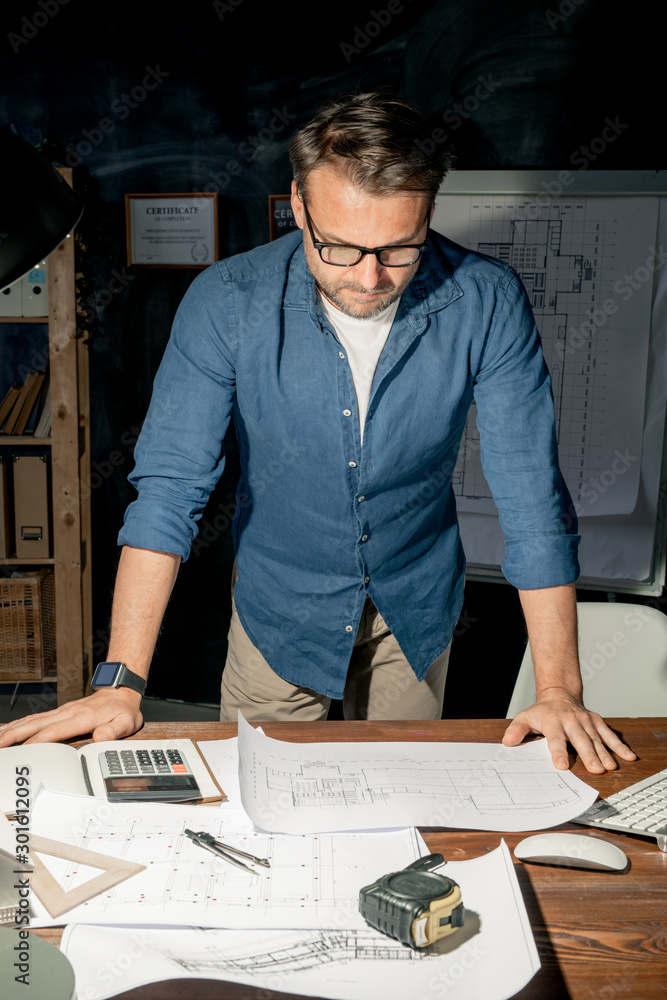 Poster mature engineer in casualwear looking at sketch on paper while working overtime