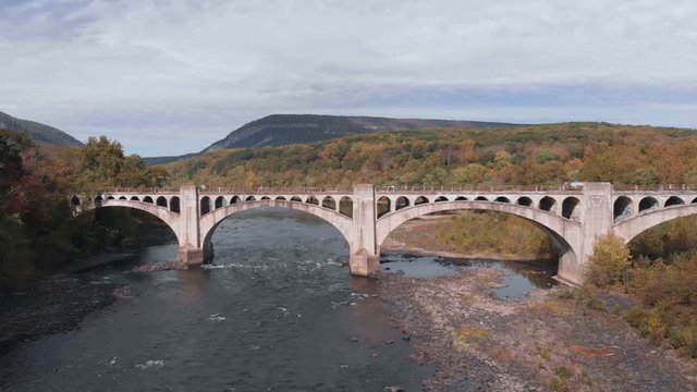Peaceful autumn afternoon flying over a shallow river flowing over rocks. Pedestrian bridge with arches over a rural river. Colorful foliage in the Pennsylvania countryside.