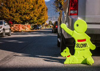Yellow slow down sign. The alert signage is features neon kid with dog, in front of cars and trees. 