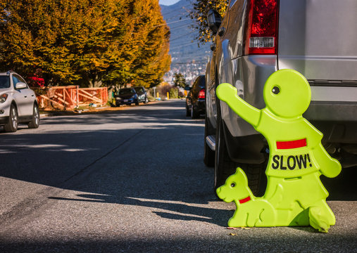 Yellow Slow Down Sign. The Alert Signage Is Features Neon Kid With Dog, In Front Of Cars And Trees. 