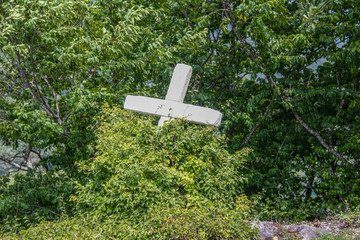 Forgotten white cross in the woods with plants growing around it - tilted and almost overgrown on hillside with more trees in background.