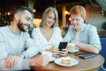 Pretty blonde girl with smartphone showing two happy guys her new photos in cafe