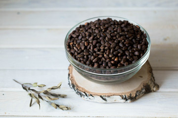 On a light wooden table on a birch saw-board there is a glass transparent deep plate full of fresh pine nuts in the shell.