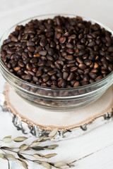 On a light wooden table on a birch saw-board there is a glass transparent deep plate full of fresh pine nuts in the shell.