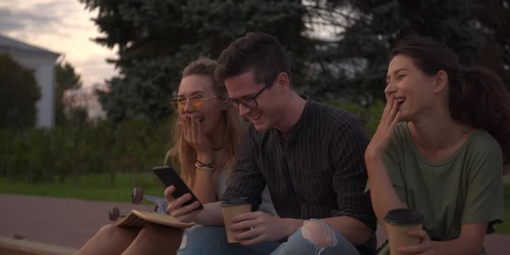 CINEMATIC SHOT of friends hipsters millennials multinational girls young women and men sitting outside drinking coffee and chatting, talking. look in phone. Slow motion