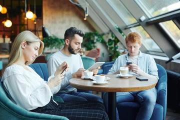 Pretty blonde girl and two guys using mobile gadgets while relaxing in cafe