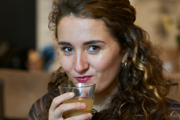 Pretty young brunette woman with curls and blonde highlights looking in the camera while holding a drink