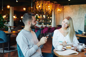 Happy blonde girl with copybook looking at young man with smartphone in cafe