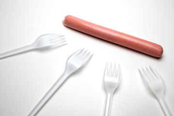 One sausage and four plastic forks on a white background. The concept of nutrition in a poor society. Hunger or lack of food. Environmental issue and the ban on plastic dishes. Copy space.