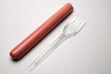 Sausage next to a plastic transparent fork on a white background. The concept of fast food, GMOs, hunger or lack of food. Environmental issue and the ban on plastic dishes. Copy space.