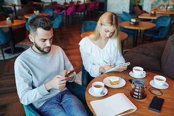 Casual guy scrolling in gadget while his girlfriend making notes or sketch