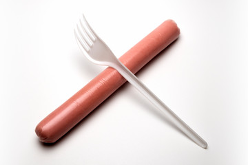 A plastic fork lies crosswise on a hot dog sausage. Ban plastic dishes. The concept of fast food, GMOs, hunger or lack of food. Ecological problem. White background. Copy space.