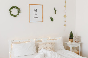 Christmas or new year winter home interior decor. Holiday decorated room. White stylish cozy scandinavian bedroom with little christmas tree, coniferous twigs or pine branches and led garland lights.