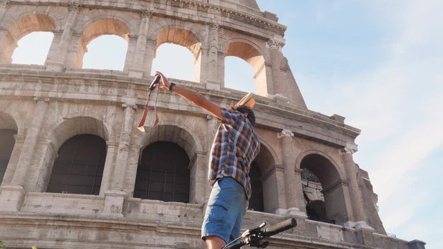 Happy young man tourist with bike wearing shirt tanding on a column taking funny pictures and selfies at colosseum in Rome, Italy at sunrise.