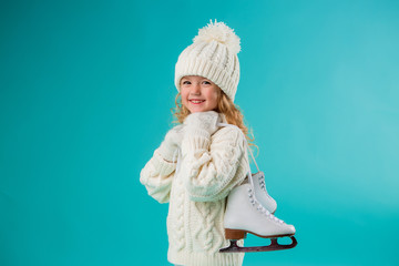 little blonde girl smiling in a winter white hat and sweater, holding skates. isolate on blue...
