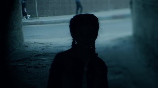 small girl silhouette with Finnish braids stands in dark gloomy gateway at backlight against people walking in street
