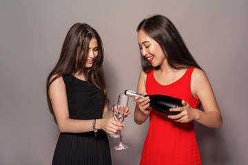 Freestyle. Young girls in dresses standing isolated on gray pouring champagne into glasses smiling happy