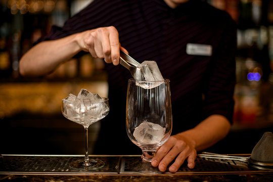 Male bartender putting an ice cube with tweezers to the empty cocktail glass