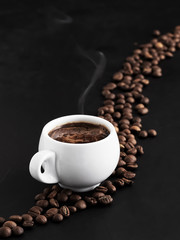White cup with fragrant espresso on a black background, steam rises above the cup. Roasted coffee beans are located around a cup of coffee and in the background. Close-up.