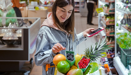 A young woman buys groceries in a supermarket with a phone in her hands.