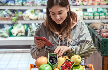 A young woman buys groceries in a supermarket with a phone in her hands.