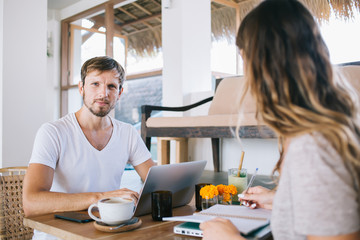 Portrait of handsome Caucasian software developer looking at camera while waiting for updating and initializing laptop computer working remotely in Indonesian coffee shop, concept of technology