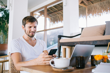 Smiling bearded man texting on cellphone during break in home office
