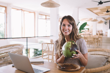 Happy female using laptop and relishing coconut juice with straw